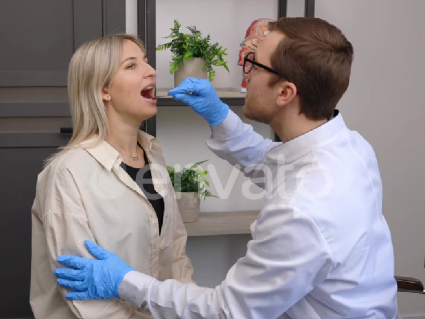 A doctor wearing gloves examines a woman’s mouth using a tongue depressor, while she sits upright with her mouth open in a calm clinical setting.